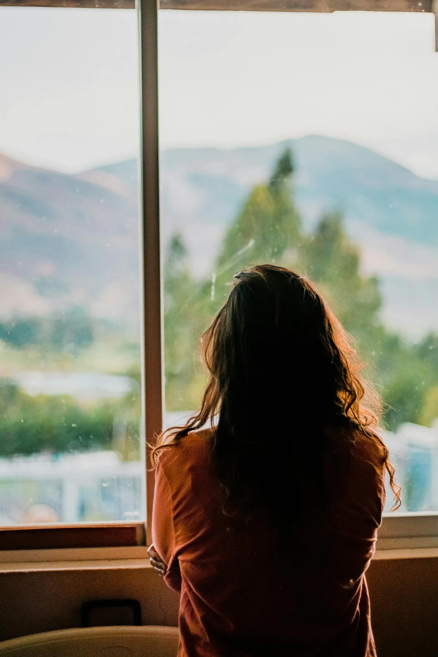 Woman standing indoors looking out through a large window toward soft green hills, reflecting quietly during a VIP Dharma Day with Nikita Thakrar in Berkshire.
